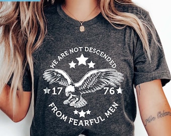 以下が含まれることがあります： 白い星と「We are not descended from fearful men 17 76」という文字が書かれた鷲のグラフィックがプリントされた濃いグレーのTシャツです。