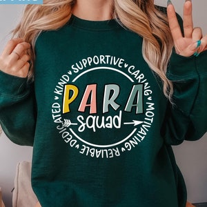 May include: A dark green sweatshirt with a white circle graphic that says "PARA squad" in colorful letters. The circle is surrounded by the words "dedicated, kind, supportive, caring, motivating, reliable".