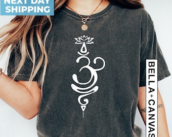 Op de afbeelding: Een grijs T-shirt met een wit Om-symbool en een lotusbloemontwerp. De tekst "BELLA + CANVAS" is zichtbaar aan de rechterkant van het shirt.