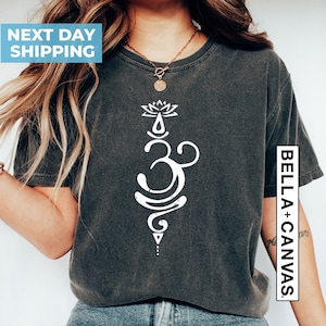 Pode incluir: Uma camiseta cinza com um s&iacute;mbolo Om branco e um design de flor de l&oacute;tus. O texto "BELLA + CANVAS" &eacute; vis&iacute;vel do lado direito da camiseta.
