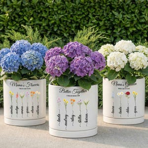 May include: Three white ceramic flower pots with colorful hydrangea blooms. The pots are labeled "Mama's Flowers", "Better Together", and "Nana's Garden". Each pot has a floral design and names.