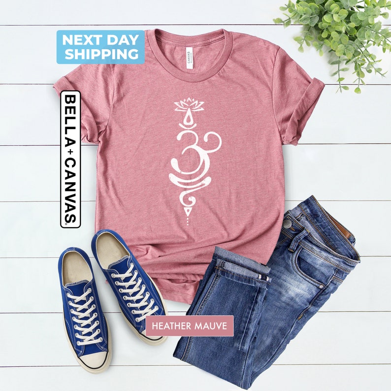 Pode incluir: Uma camiseta de cor malva desbotada com um s&iacute;mbolo Om branco e um design de flor de l&oacute;tus. A camiseta &eacute; combinada com jeans azul e t&ecirc;nis azuis.