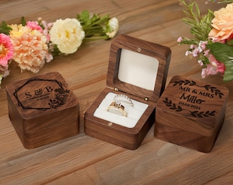 May include: Three wooden ring boxes, two closed and one open, displaying a diamond ring. The open box reveals a white interior. The closed boxes have engraved text and floral designs. One box reads "S & B" and the other reads "MR & MRS Miller 09.02.2025".