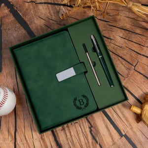 May include: A green leatherette journal and pen set in a gift box. The journal has a silver clasp and a debossed initial "B" within a laurel wreath. A matching pen and refill are included. The set is presented on a wooden surface.
