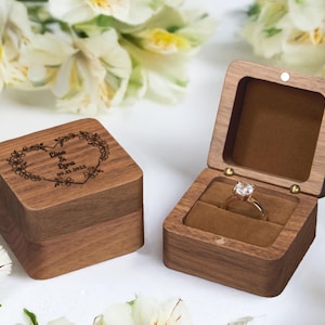 May include: Two wooden ring boxes. One closed box features a heart-shaped floral design with the names "Elisa & Eyra" and the date "09.11.2023". The other box is open, revealing a diamond ring set on a brown velvet interior.