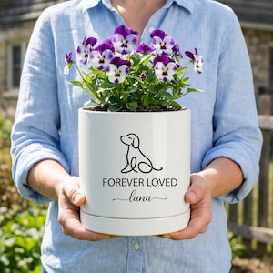 May include: A white ceramic planter with a black line drawing of a dog and the text "FOREVER LOVED luna". The planter holds purple and white pansies. The planter is held by a person wearing a blue shirt.
