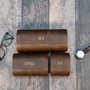 May include: Three brown leather watch cases with gold lettering. The cases are rectangular and have a roll-up design. The largest case has "RV" on it, the medium case has "HND", and the smallest case has "DF".