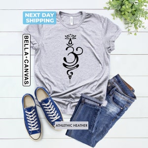 Pode incluir: Camiseta cinza com um s&iacute;mbolo Om preto e um design de flor de l&oacute;tus. O texto "ATHLETIC HEATHER" est&aacute; impresso na parte inferior da camiseta.