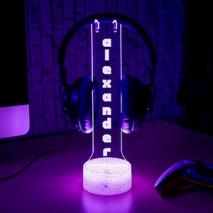 May include: A purple-lit headphone stand with the word "diexander" vertically displayed. The stand holds a pair of black headphones. The base of the stand has a cracked pattern design. A game controller is visible in the foreground.