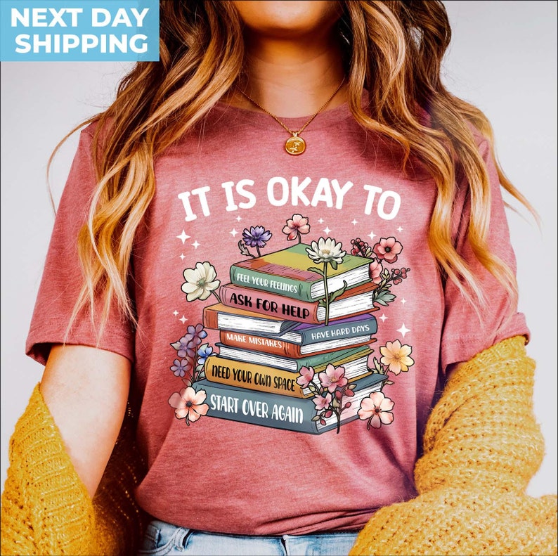 K&ouml;nnte beinhalten: Ein pinkfarbenes T-Shirt mit dem Text "It is okay to" und einem Stapel B&uuml;cher mit S&auml;tzen wie "Feel your feelings", "Ask for help", "Make mistakes", "Have hard days", "Need your own space" und "Start over again".
