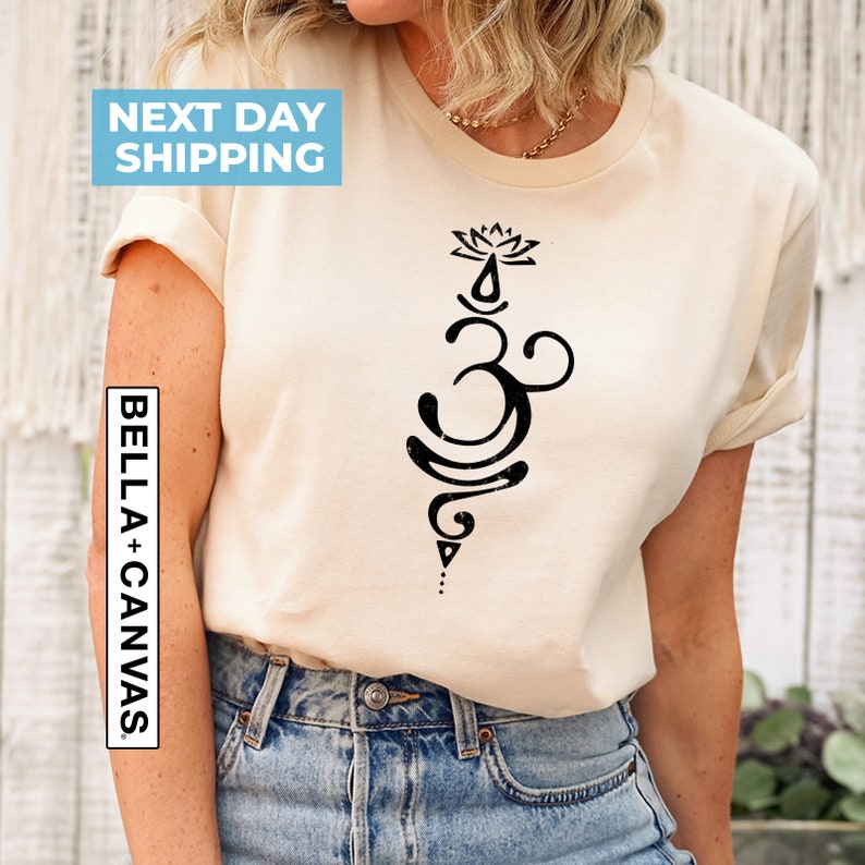 Pode incluir: Uma camiseta bege com um s&iacute;mbolo Om preto e um design de flor de l&oacute;tus. O texto "BELLA + CANVAS" est&aacute; impresso no lado esquerdo da camiseta.