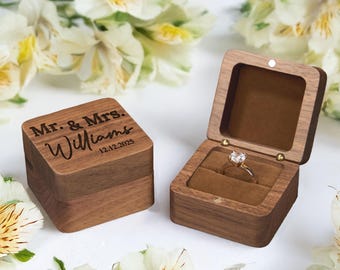 May include: A wooden ring box with the inscription "Mr. & Mrs. Williams 12.12.2025" on the lid. The box is open, revealing a gold ring with a clear gemstone set in a brown velvet interior. The box is made of dark wood.