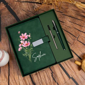 May include: A dark green gift set containing a notebook and a matching pen. The notebook cover features a pink floral illustration and the name Sophia in white script. A silver metal clasp secures the notebook, and a pen is included in the box.