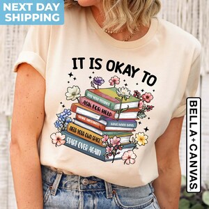 K&ouml;nnte beinhalten: Ein beiges T-Shirt mit dem Text "It is okay to" und einem Stapel B&uuml;cher mit S&auml;tzen wie "Feel your feelings", "Ask for help", "Make mistakes", "Have hard days", "Need your own space" und "Start over again".
