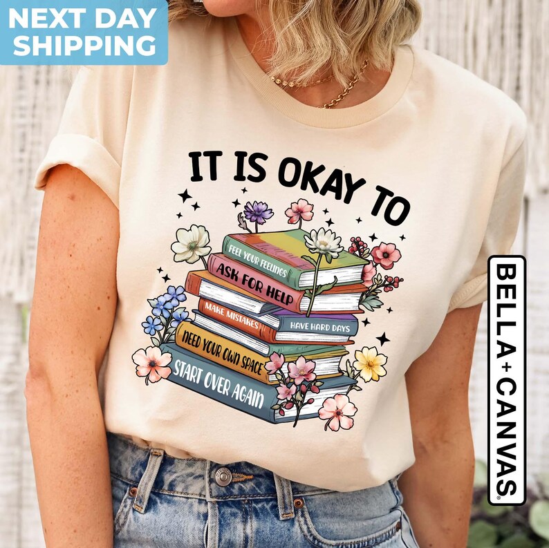 K&ouml;nnte beinhalten: Ein beiges T-Shirt mit dem Text "It is okay to" und einem Stapel B&uuml;cher mit S&auml;tzen wie "Feel your feelings", "Ask for help", "Make mistakes", "Have hard days", "Need your own space" und "Start over again".