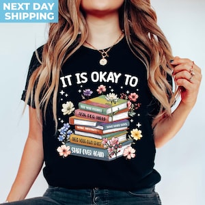 K&ouml;nnte beinhalten: Schwarzes T-Shirt mit einem Stapel bunter B&uuml;cher mit dem Text "Es ist okay zu" und dann aufgelistet "Deine Gef&uuml;hle f&uuml;hlen, Hilfe holen, Fehler machen, Schwierige Tage haben, Deinen eigenen Raum brauchen, Von vorne anfangen".