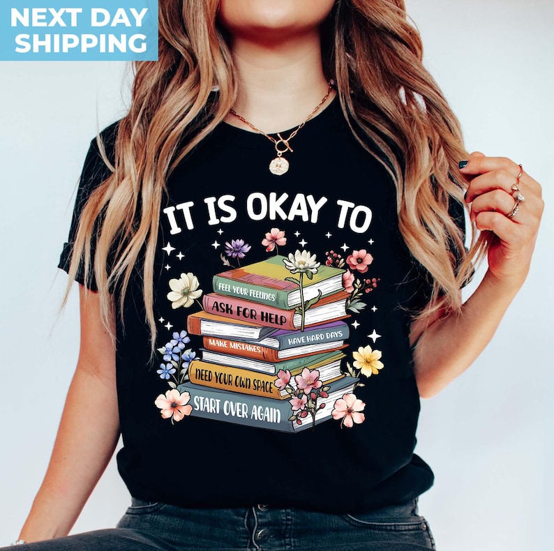 K&ouml;nnte beinhalten: Schwarzes T-Shirt mit einem Stapel bunter B&uuml;cher mit dem Text "Es ist okay zu" und dann aufgelistet "Deine Gef&uuml;hle f&uuml;hlen, Hilfe holen, Fehler machen, Schwierige Tage haben, Deinen eigenen Raum brauchen, Von vorne anfangen".