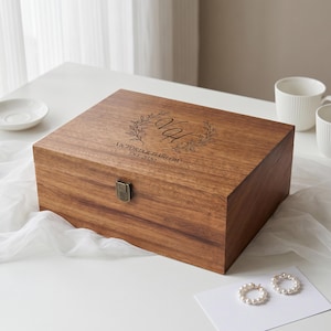 May include: A rectangular wooden keepsake box with a dark brown finish and a metal latch. The lid features an engraved design with the initials VH surrounded by a leaf wreath, and the text VICTORIA & HARLOW EST. 2020.