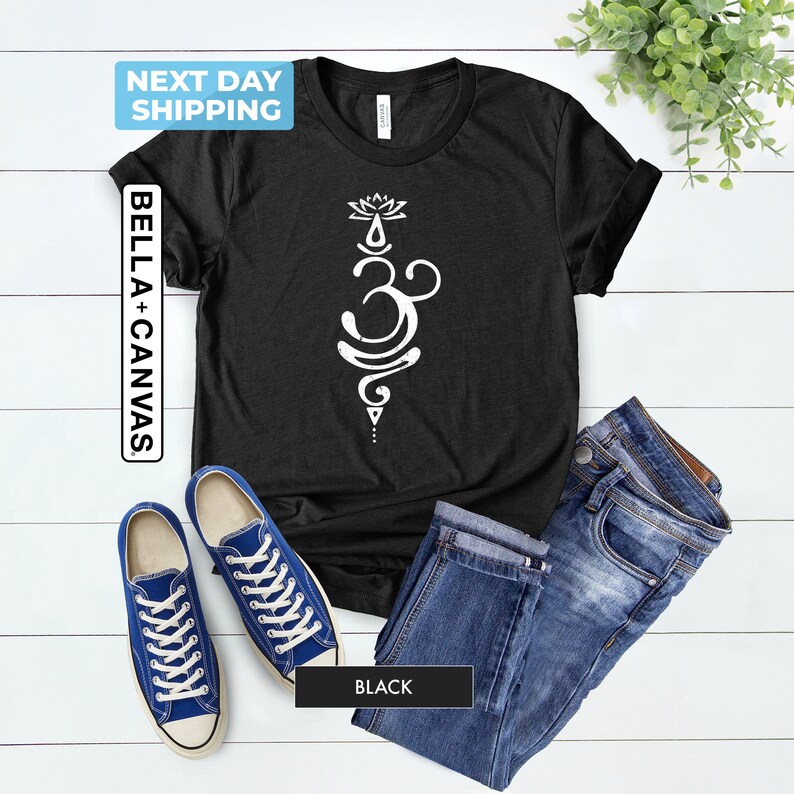 Pode incluir: Camiseta preta com um s&iacute;mbolo Om branco e um design de flor de l&oacute;tus. A camiseta est&aacute; colocada sobre uma superf&iacute;cie de madeira branca com um par de t&ecirc;nis azuis e uma cal&ccedil;a jeans azul.