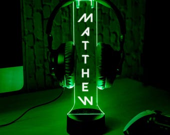 May include: A personalized headphone stand illuminated with green light. The stand displays the name "MATTHEW" vertically in white lettering. Black headphones rest on the stand, and a gaming controller is visible in the foreground.