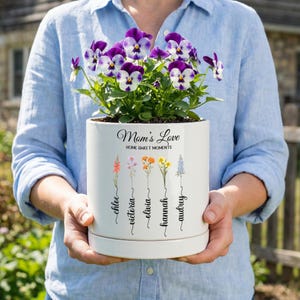 May include: A white ceramic flower pot with the text "Mom's Love" and "Home Sweet Moments". The pot features illustrations of flowers and names: Chloe, Victoria, Olivia, Hannah, and Audrey. The pot contains purple and white pansies.