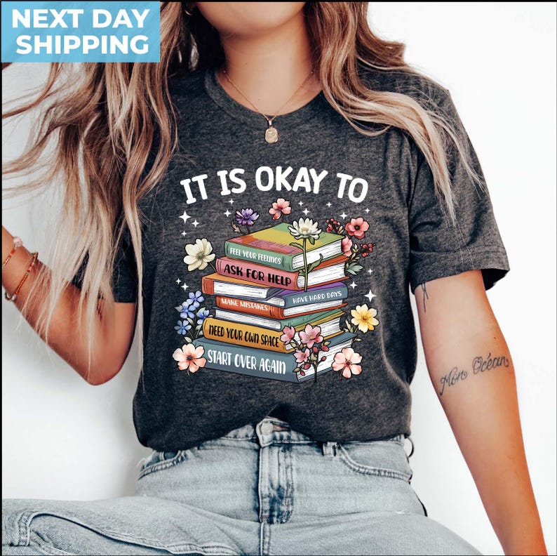 K&ouml;nnte beinhalten: Ein dunkelgraues T-Shirt mit dem Text "It is okay to" und einem Stapel B&uuml;cher mit dem Text "Feel your feelings", "Ask for help", "Make mistakes", "Have hard days", "Need your own space" und "Start over again". Die B&uuml;cher sind von bunten Blumen umgeben.