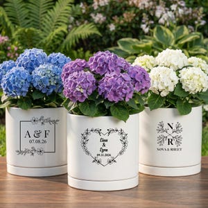 May include: Three white ceramic planters with vibrant hydrangea blooms in blue, purple, and white. Each pot features a unique black floral design with initials and dates, perfect for home decor or special events.