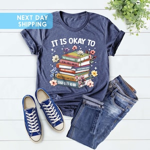 K&ouml;nnte beinhalten: Ein marineblaues T-Shirt mit einem Stapel B&uuml;cher und Blumen. Der Text auf dem Shirt lautet "It is okay to" und "Ask for help" und "Start over again".