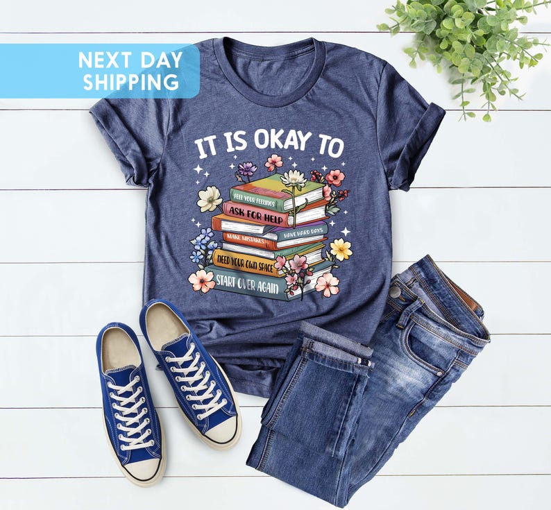 K&ouml;nnte beinhalten: Ein marineblaues T-Shirt mit einem Stapel B&uuml;cher und Blumen. Der Text auf dem Shirt lautet "It is okay to" und "Ask for help" und "Start over again".
