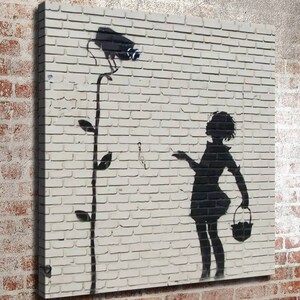 May include: A black and white stencil art print of a girl holding a basket and a flower with a security camera on top. The artwork is on a white brick wall background.