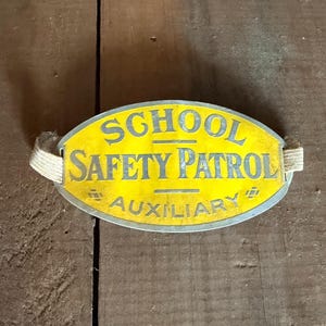 May include: Vintage School Safety Patrol badge. The badge is oval-shaped with a yellow background and a silver border. The text "SCHOOL SAFETY PATROL AUXILIARY" is printed in blue. The badge has two white fabric straps attached to the sides.