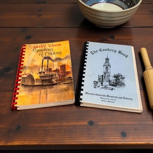 May include: Two spiral-bound cookbooks, one titled The New Saint Louis Symphony of Cooking with a riverboat illustration, and one titled The Cookery Book from the Winston Churchill Memorial and Library, alongside a wooden rolling pin and a ceramic bowl.
