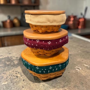 May include: A stack of three hexagonal woven baskets with light wood lids. The top basket has a tan fabric liner, the middle basket features a burgundy fabric liner with a white floral pattern, and the bottom basket has a dark green floral liner.