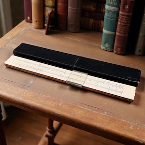 May include: A vintage slide rule with a black case, resting on a wooden surface. The slide rule is beige with black markings and a metal slider. The case is rectangular and open, revealing the slide rule. Books are in the background.