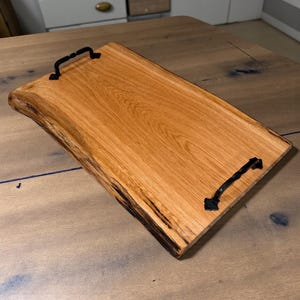 May include: A rectangular wooden serving tray with a natural edge and two black metal handles. The tray is made of light brown wood with visible grain and knots. The handles are attached at each end.