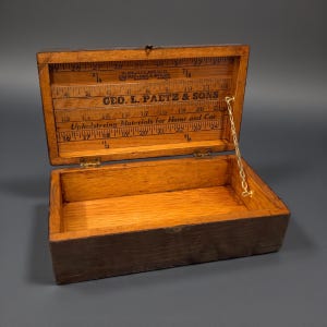 May include: A rectangular wooden box with an open lid featuring a printed ruler and the text Geo. L. Paetz & Sons Upholstering Materials for Home and Car. The interior is natural wood, and a small metal chain connects the lid to the base.