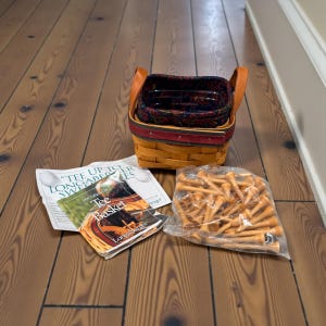 May include: A woven basket with leather handles, a red band, and a patterned interior. Beside it are a brochure with the text "Tee Basket" and a clear bag filled with wooden golf tees. The items are on a wooden floor.