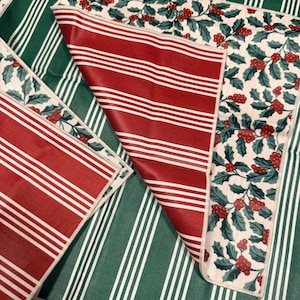 May include: A reversible fabric placemat featuring a festive holiday pattern on one side with green holly leaves and red berries on a cream background, and a classic red and white striped pattern on the reverse side with finished edges.
