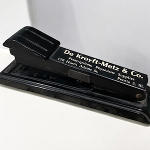 May include: Black metal stapler with a gold inscription that reads "De Kroyft-Metz & Co. Prescriptions, Physicians Supplies 126 North Adams St. Peoria 2, Ill."