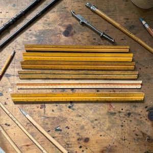 May include: A collection of vintage yellow wooden rulers with black markings, arranged on a wooden surface. Pencils and other drafting tools are scattered around the rulers. The rulers appear to be in good condition.