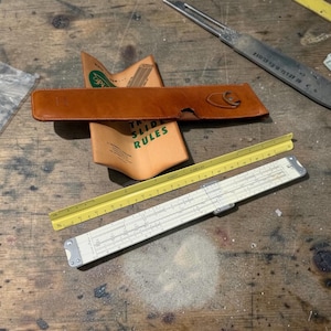 May include: A collection of vintage drafting tools on a wooden surface. The items include a leather case, a slide rule, a yellow ruler, and a booklet with the text "Slide Rules". The slide rule is white with black markings.