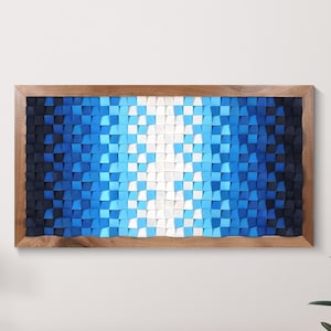 Puede incluir: Una pieza de arte de pared de madera con un patrón geométrico azul, blanco y negro. El arte está enmarcado en un marco de madera oscura y está colgado en una pared blanca sobre una cómoda de madera.