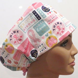 May include: A white surgical scrub hat with a pink ribbon pattern. The hat has various phrases printed on it, including "Think Pink", "Brave & Strong", "Peace Love", "You Are A Warrior", and "Support Admire".