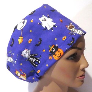 May include: A blue Halloween-themed scrub cap featuring cartoon cats dressed as a ghost, mummy, and skeleton, along with bats, candy corn, and pumpkins. The cap is made of fabric and is designed to be worn on the head.