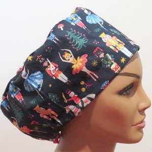 May include: A black scrub hat with a festive Nutcracker and ballerina print. The hat features a variety of colourful characters, including a Nutcracker soldier, a ballerina, and a Christmas tree.