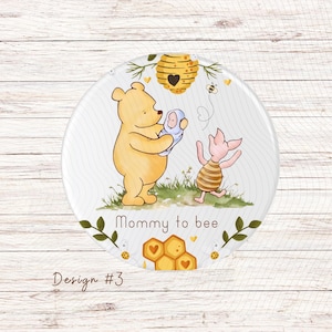 May include: A round, white, wooden-textured sign with a Winnie the Pooh and Piglet illustration. Winnie the Pooh is holding a baby in a blue blanket. Piglet is wearing a yellow and black striped shirt. The sign has a honeycomb design and the text "Mommy to bee".