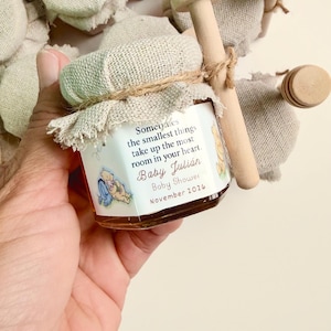 May include: A small glass jar filled with honey, topped with a beige fabric cover tied with twine and a wooden honey dipper. The label reads: Sometimes the smallest things take up the most room in your heart. Baby Julián Baby Shower November 2026.