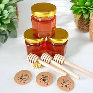 May include: Three small glass jars filled with honey, stacked on top of each other. The jars have gold lids and are labeled with "Thank You" and hearts.  Three wooden honey dippers and three round brown paper tags with the text "Thank You so much" are in front of the jars.