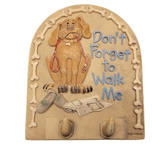 Könnte beinhalten: Ein beigefarbener Keramikhaken in Form eines Hundebeins mit einem Comic-Hund darauf. Der Hund ist braun mit einem roten Halsband und einer Leine. Der Haken hat zwei Haken unten. Der Text auf dem Haken lautet "Vergiss nicht, mich Gassi zu führen".