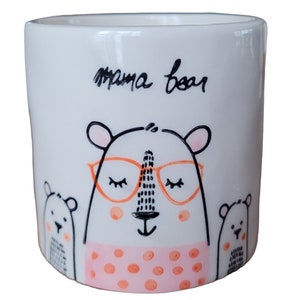 Puede incluir: Taza de cerámica blanca con una ilustración en negro y naranja de una mamá oso con gafas y dos ositos. La taza dice "mama bear" en cursiva negra.